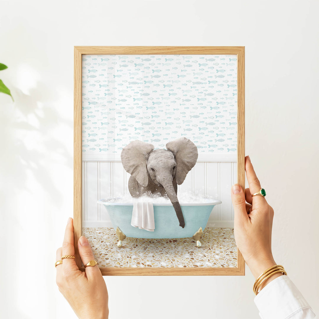A person's hands are holding a framed picture of a baby elephant sitting in a bathtub filled with water and bubbles.