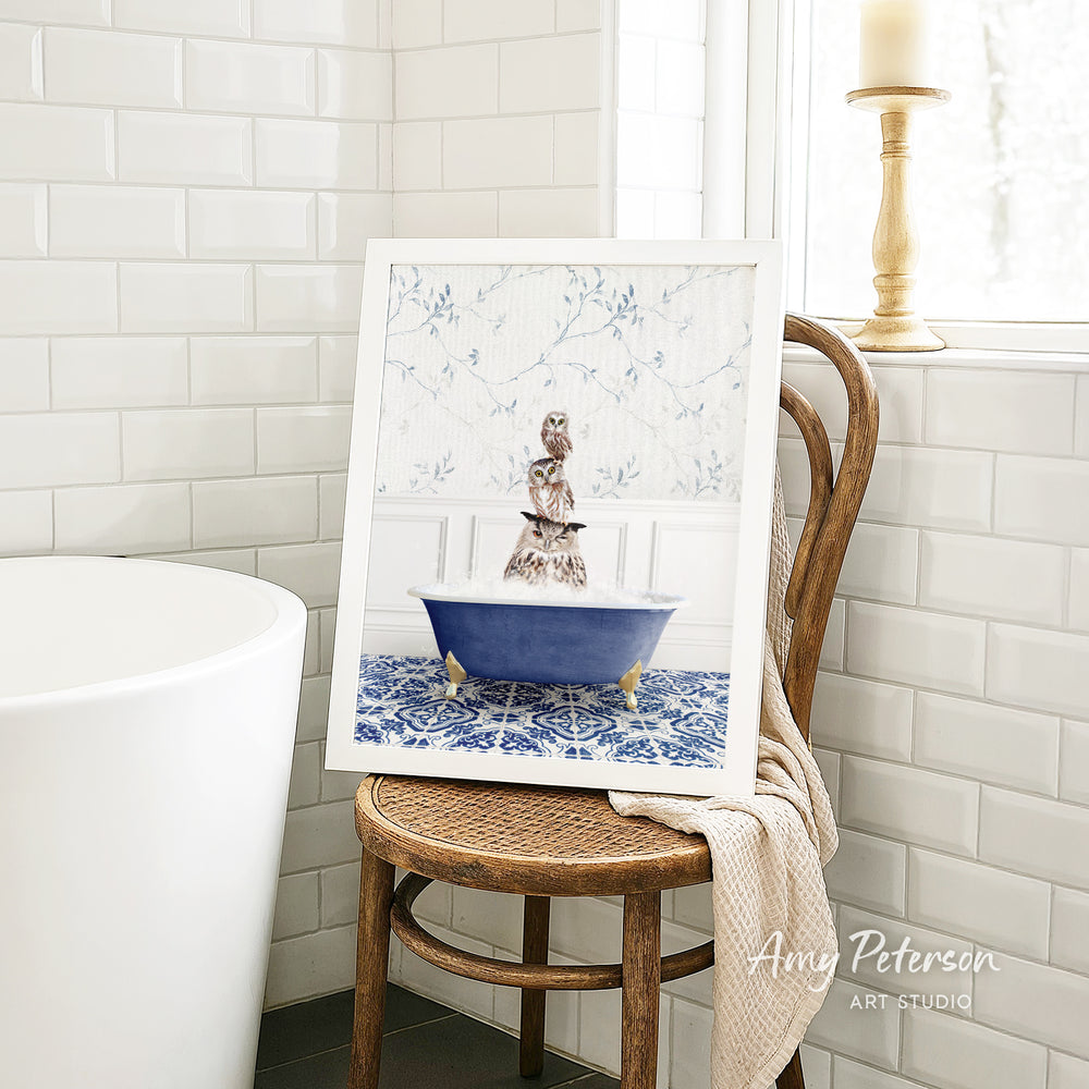 A framed image of a cat sitting in a blue bathtub, surrounded by a floral pattern, is displayed on a wooden chair in a bathroom setting.