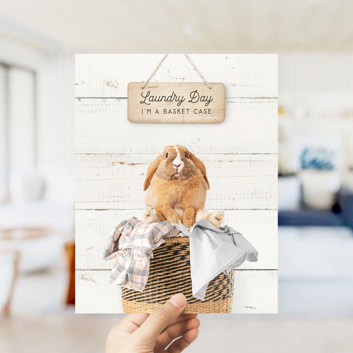 A hand holding a card with a picture of a rabbit sitting in a basket filled with clothes, with a wooden sign above it that says "Laundry Day I'm a basket case".