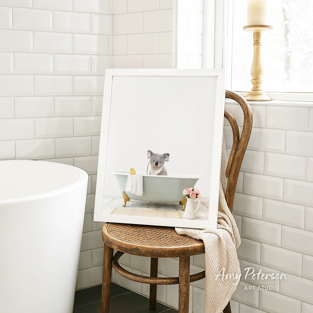 A framed image of a koala bear sitting in a bathtub, with a vase of flowers and a towel nearby, is displayed on a wooden chair in a bathroom setting.