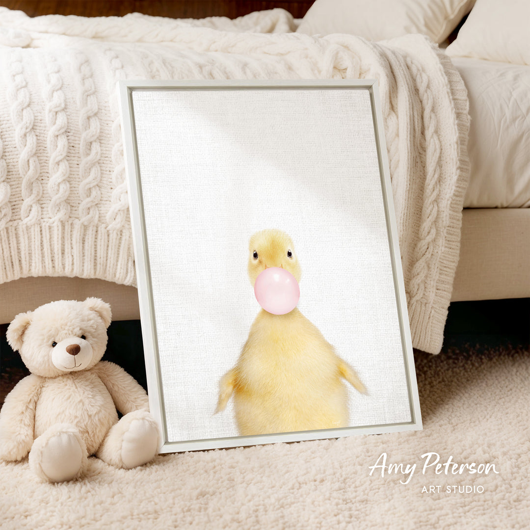 A framed image of a yellow duck blowing a bubble gum bubble is displayed on a white bedspread, accompanied by a teddy bear.