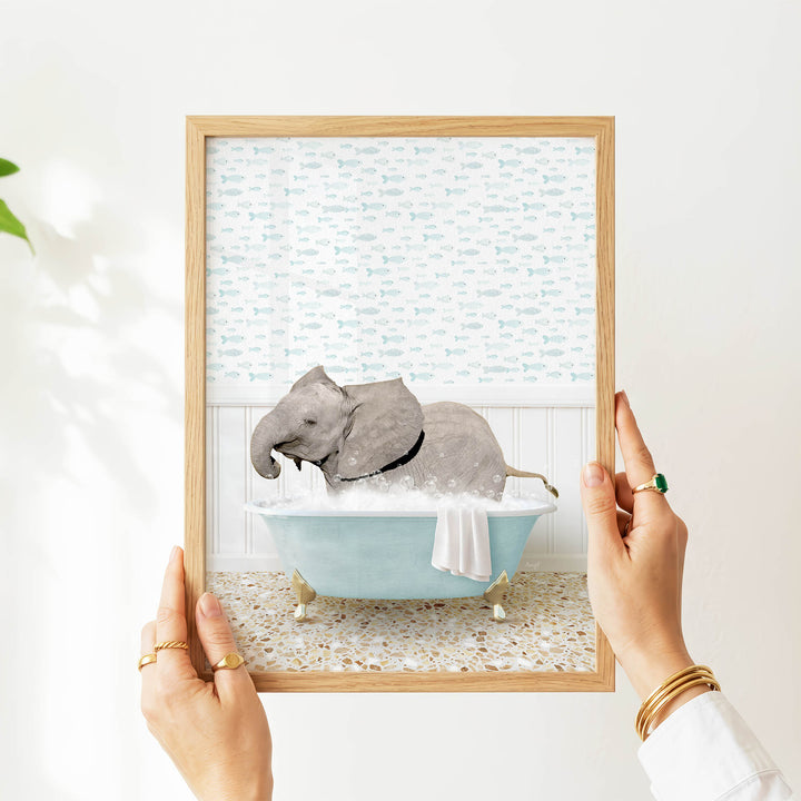 A person's hands are holding a framed picture of an elephant sitting in a bathtub filled with water, surrounded by a pattern of fish.