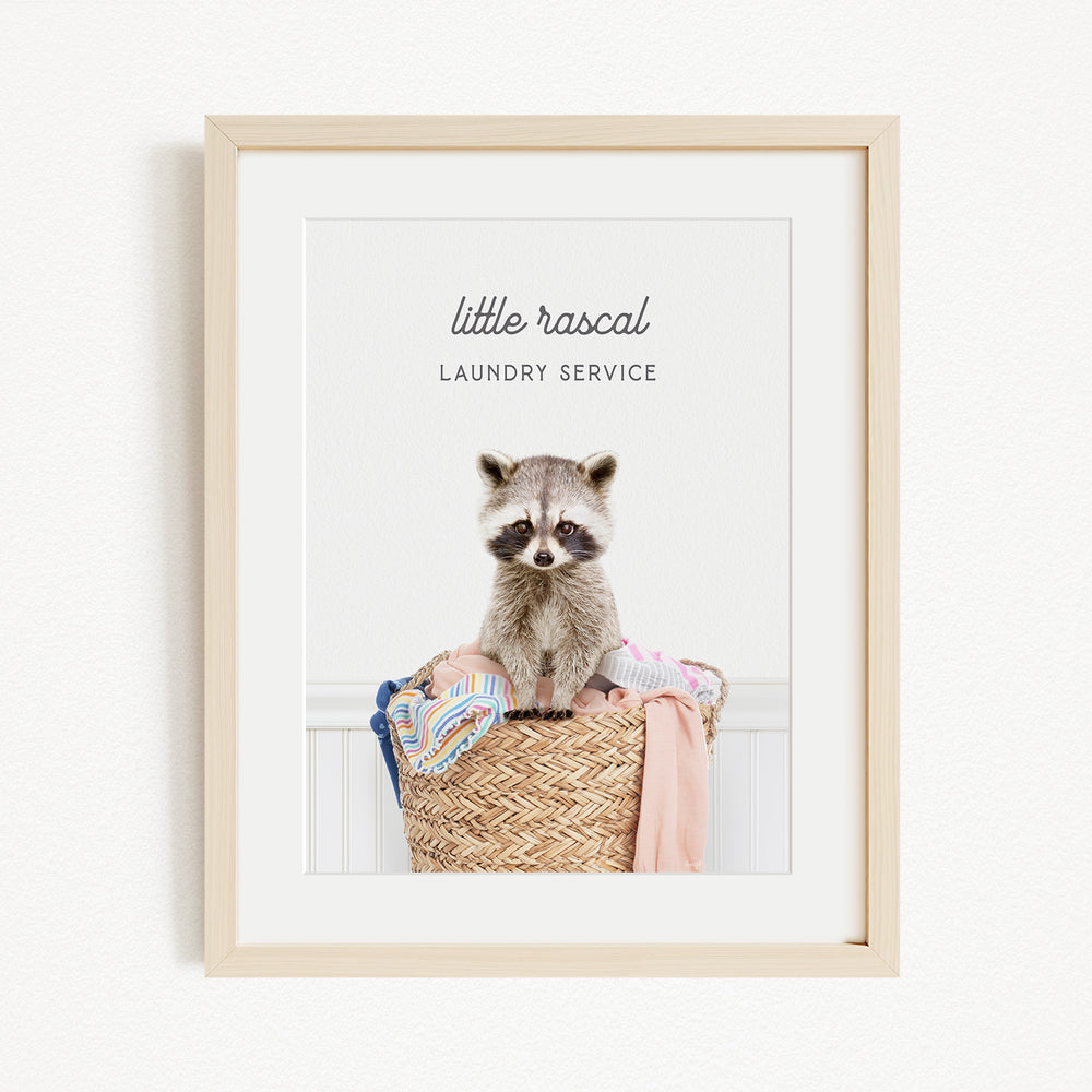 A raccoon sitting in a woven laundry basket, with the text "little rascal Laundry Service" displayed above the raccoon.