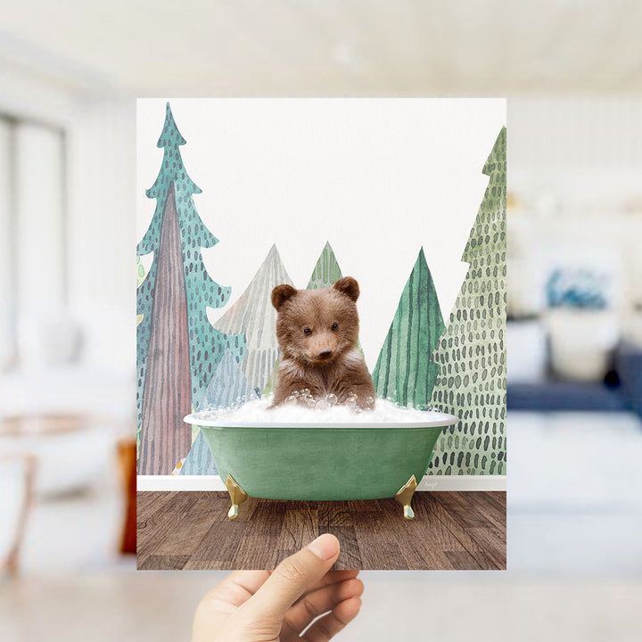 A hand is holding up a framed image of a small brown bear sitting in a green bathtub, surrounded by a winter landscape with trees and snow.