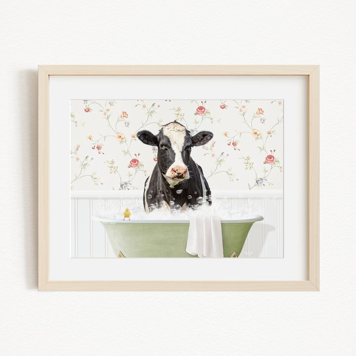 A black and white cow is sitting in a green bathtub filled with bubbles, with a floral wallpaper in the background.