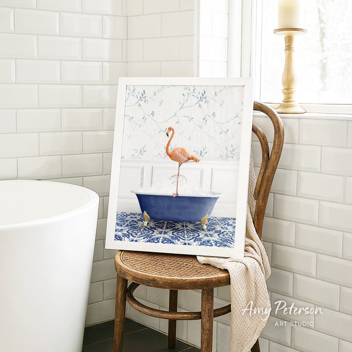A framed image of a pink flamingo standing in a blue bathtub is displayed on a wooden chair in a bathroom setting.