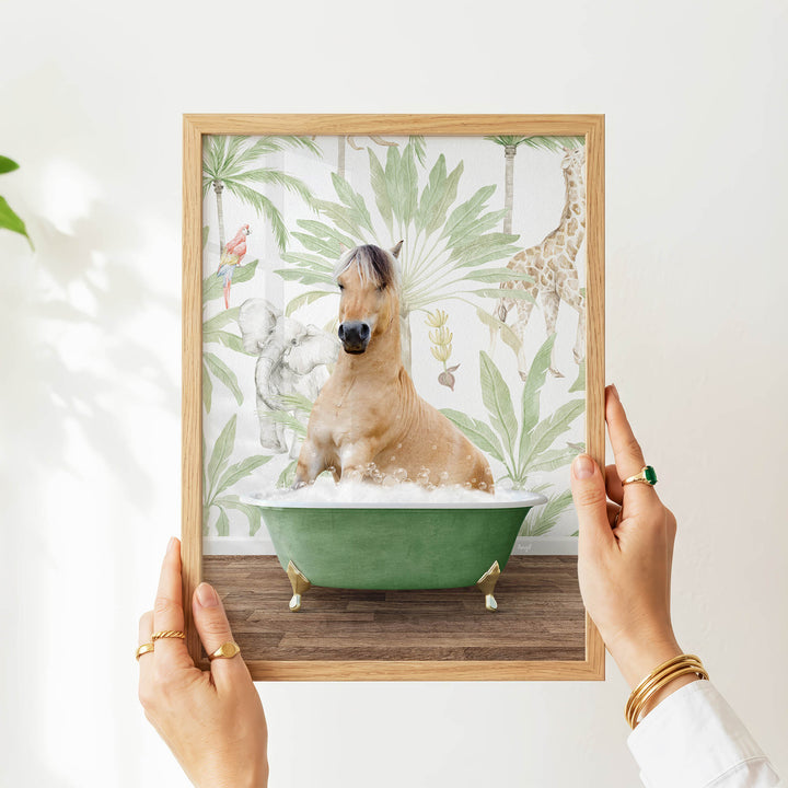 A person's hands are holding a framed picture of a horse sitting in a bathtub, surrounded by tropical foliage.