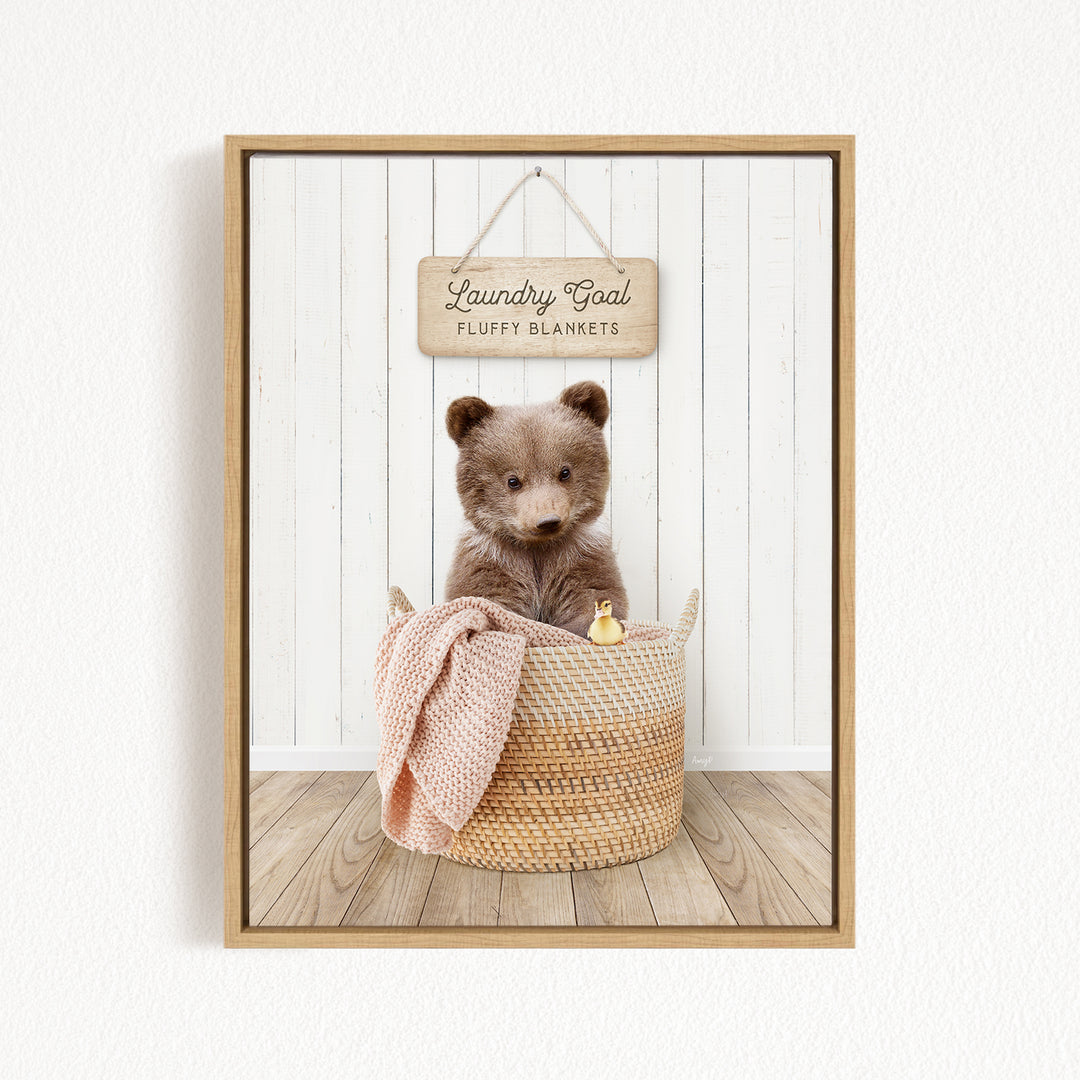 A brown teddy bear sits in a woven basket filled with blankets, with a wooden sign above that reads "Laundry Goal Fluffy Blankets".
