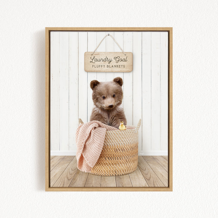 A brown teddy bear sits in a woven basket filled with blankets, with a wooden sign above that reads "Laundry Goal Fluffy Blankets".