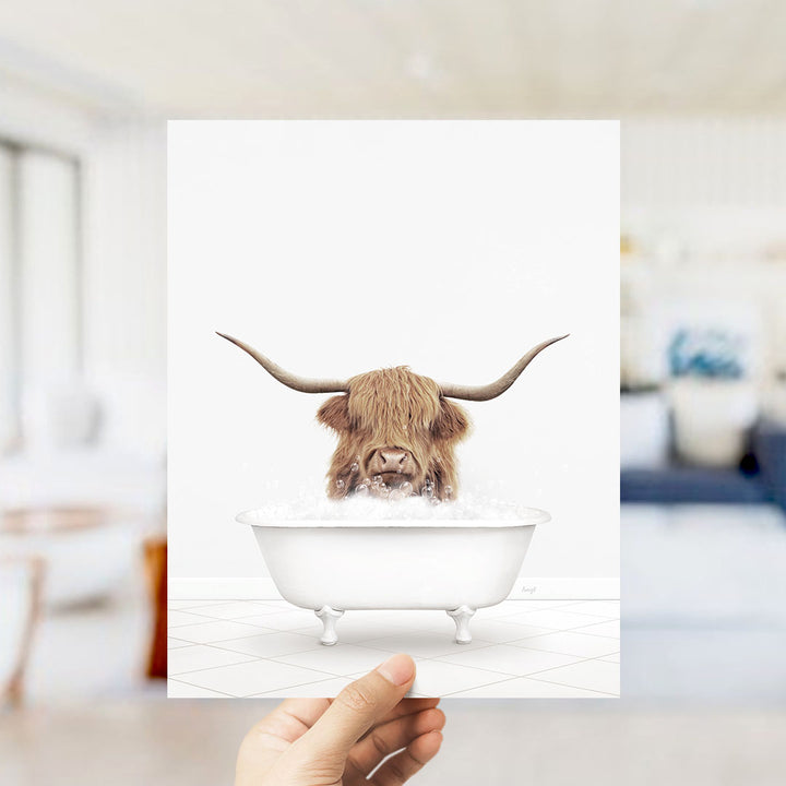 A hand is holding up a framed image of a Highland cow with large horns sitting in a bathtub filled with water.