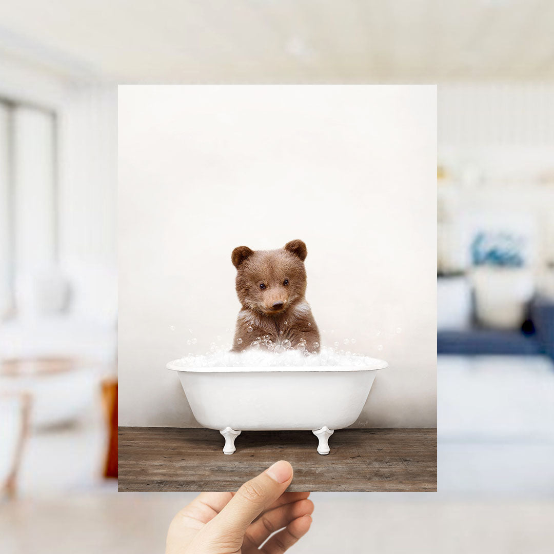 A hand holds up a framed image of a small brown bear sitting in a white bathtub filled with bubbles.
