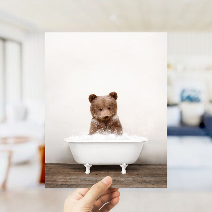 A hand holds up a framed image of a small brown bear sitting in a white bathtub filled with bubbles.