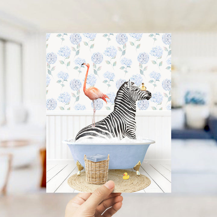 A hand holds up a framed image depicting a zebra sitting in a bathtub, with a pink flamingo standing on its back and a yellow rubber duck floating nearby.