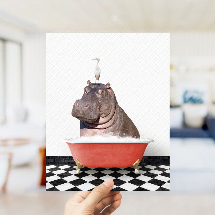 A hand holds up a framed image depicting a hippopotamus sitting in a red bathtub, with a seagull perched on its head.