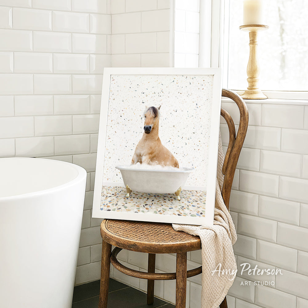 A framed image of a horse sitting in a bathtub, surrounded by a white and beige color scheme, is displayed on a wooden chair in a bathroom setting.