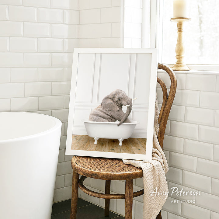 A framed photograph of a gray elephant sitting in a white bathtub, with a wooden chair and a white tiled wall in the background.