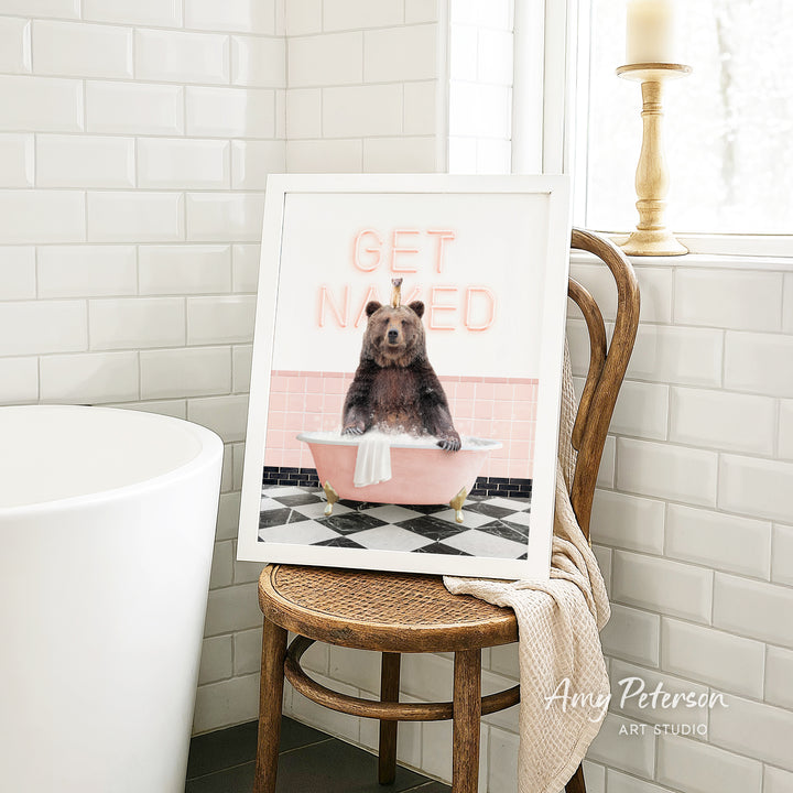 A framed image of a bear sitting in a bathtub with the text "GET NAKED" on it, placed on a wooden chair in a bathroom setting.