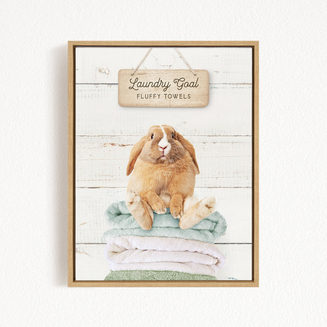 A framed image of a fluffy towel with a rabbit sitting on top of it, against a white wooden background.