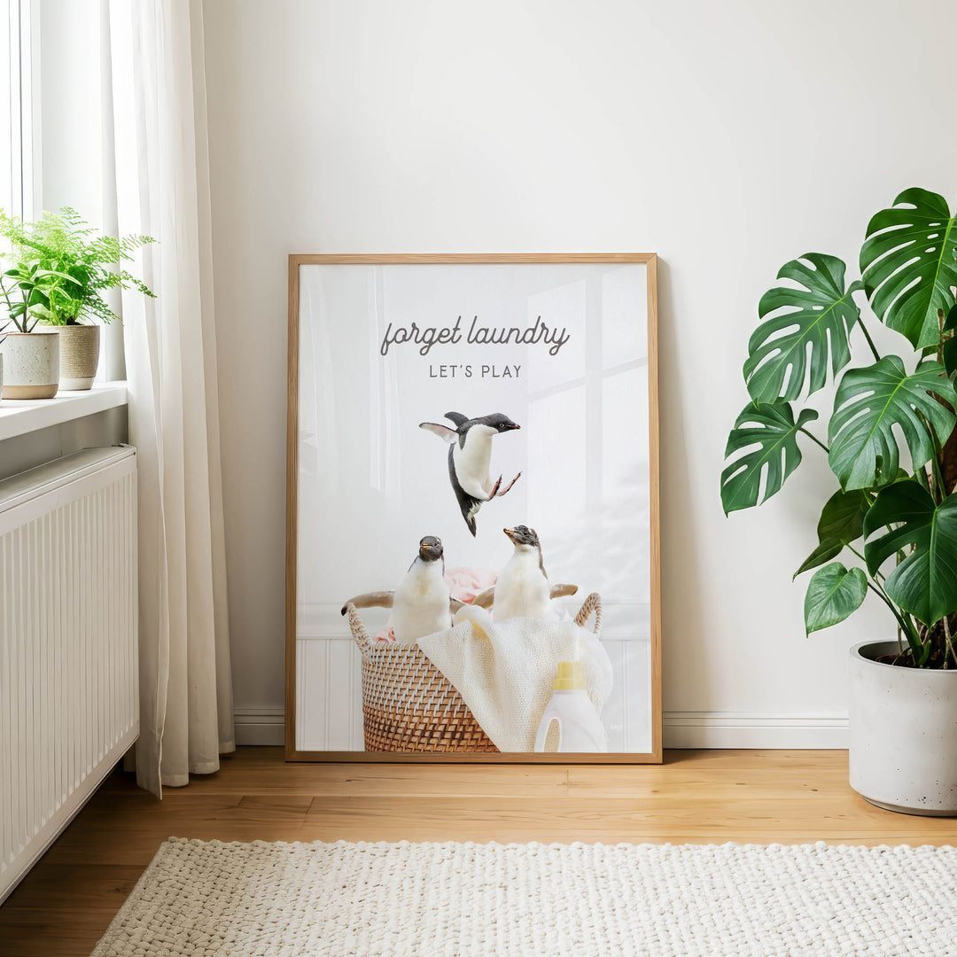 A framed poster with a penguin image and the text "Forget laundry, let's play" is displayed on a wall in a room with a rug and potted plants.