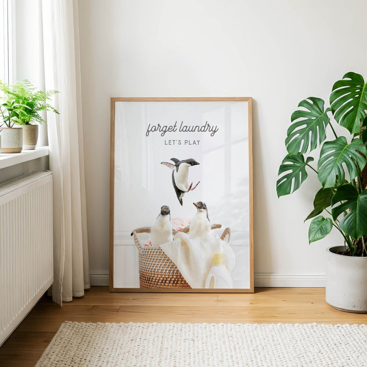 A framed poster with a penguin image and the text "Forget laundry, let's play" is displayed on a wall in a room with a rug and potted plants.