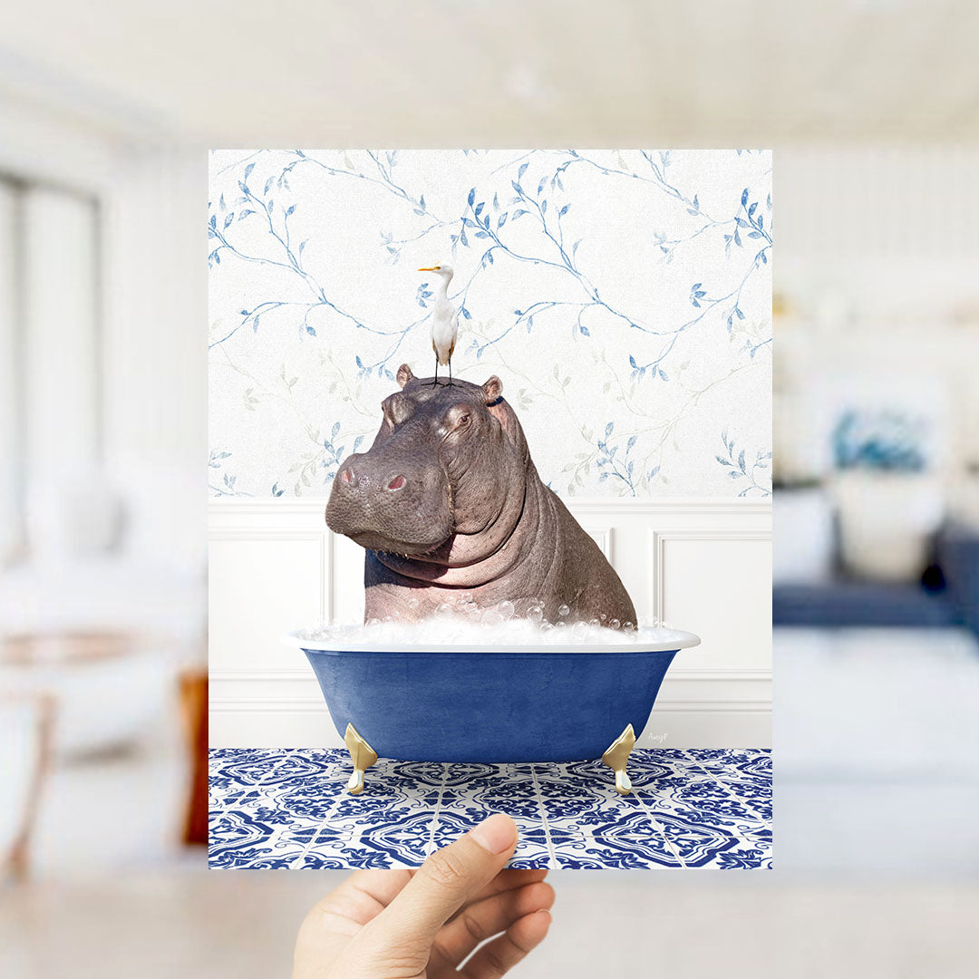 A hand holds up a framed image of a hippopotamus sitting in a blue bathtub, with a bird perched on its head.