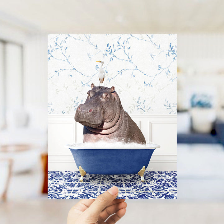 A hand holds up a framed image of a hippopotamus sitting in a blue bathtub, with a bird perched on its head.
