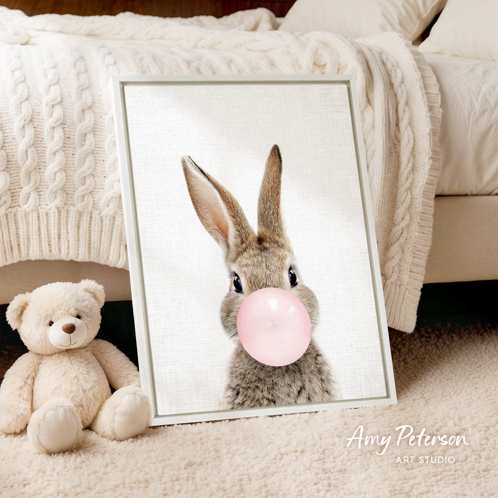 A framed image of a rabbit blowing a bubble with a pink bubble gum bubble, accompanied by a teddy bear on a bed.