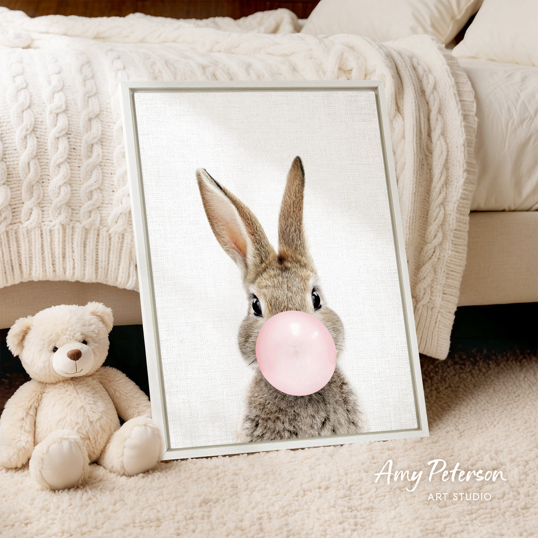 A framed image of a rabbit blowing a bubble with a pink bubble gum bubble, accompanied by a teddy bear on a bed.