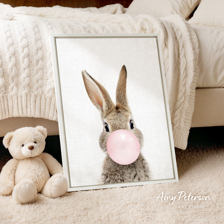 A framed image of a rabbit blowing a bubble with a pink bubble gum bubble, accompanied by a teddy bear on a bed.