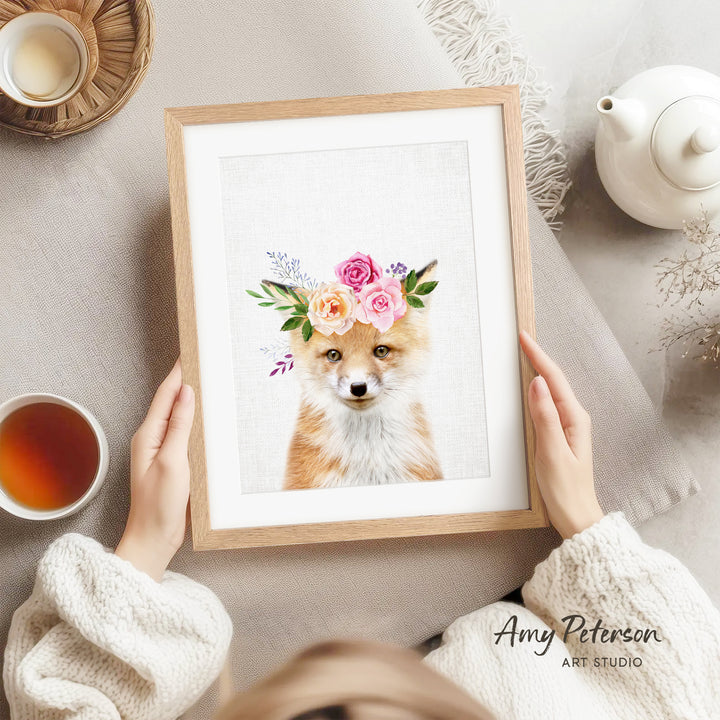 A person is holding a framed picture of a fox wearing a floral crown, with a teapot, a cup of tea, and a bowl of tea nearby.