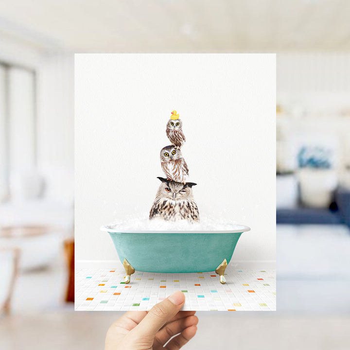 A hand holds up a framed image depicting three owls sitting in a bathtub filled with bubbles.