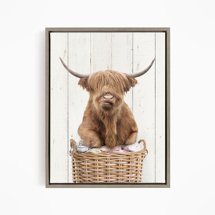 A Highland cow with shaggy brown hair sits in a wicker basket against a white wooden background.