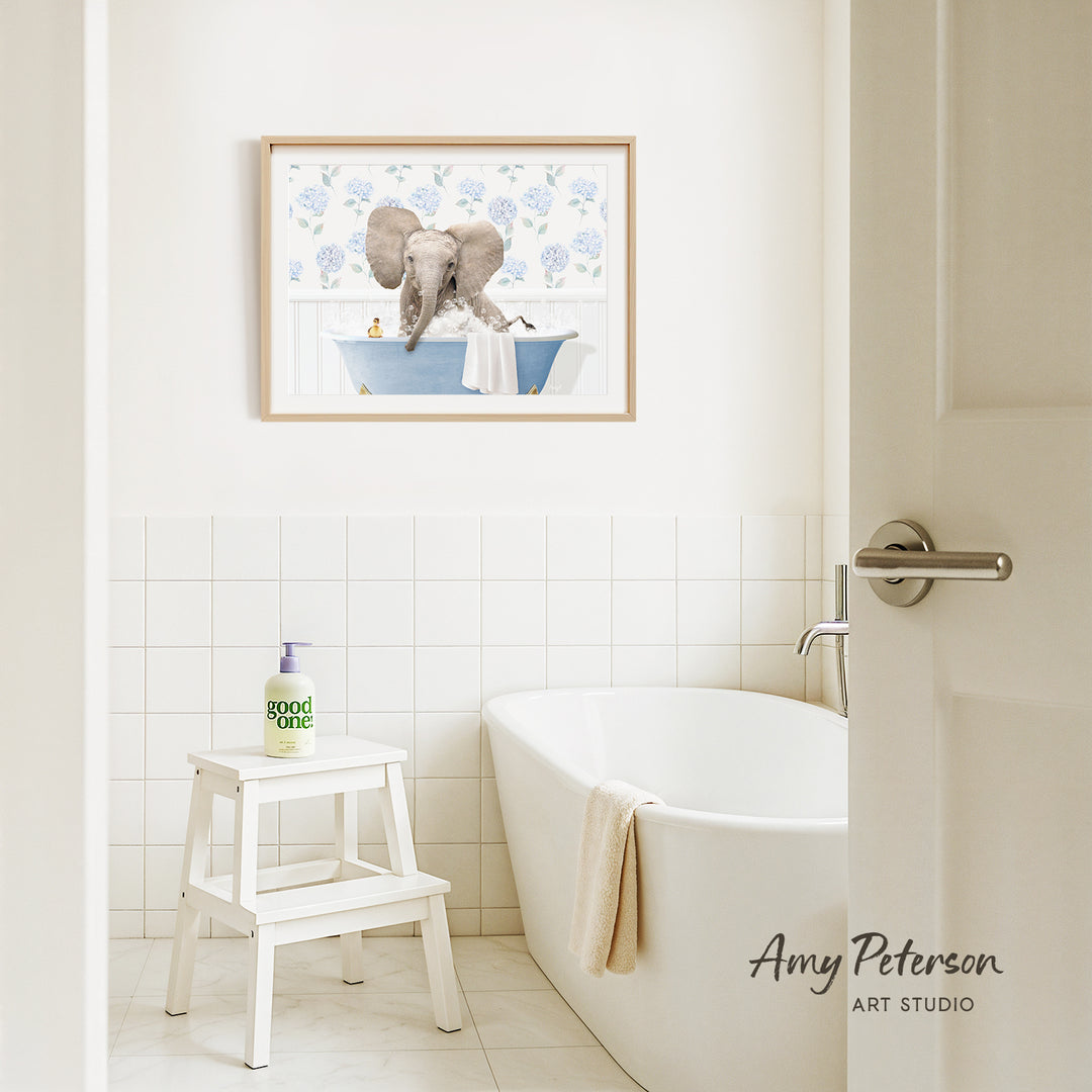 A framed image of an elephant sitting in a bathtub is hanging on a white tiled wall above a white bathtub in a bathroom.