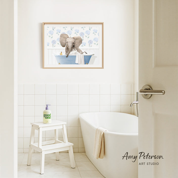 A framed image of an elephant sitting in a bathtub is hanging on a white tiled wall above a white bathtub in a bathroom.