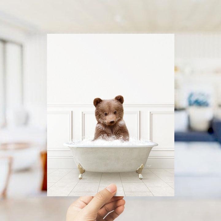 A hand holds up a framed image of a small brown bear sitting in a white bathtub filled with bubbles.