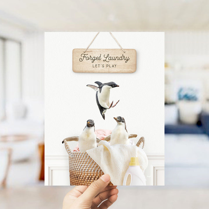 A hand holds up a framed image of three penguins in a basket, with a wooden sign above them that says "Forget Laundry Let's Play".