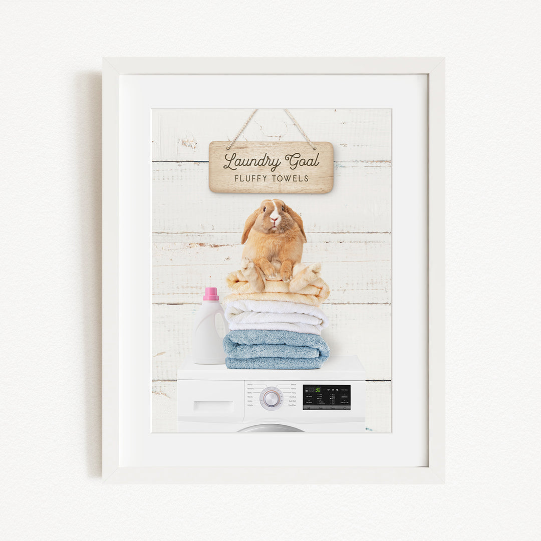 A framed image depicting a rabbit sitting on a stack of fluffy towels, with a sign above that reads "Laundry goal Fluffy towels".