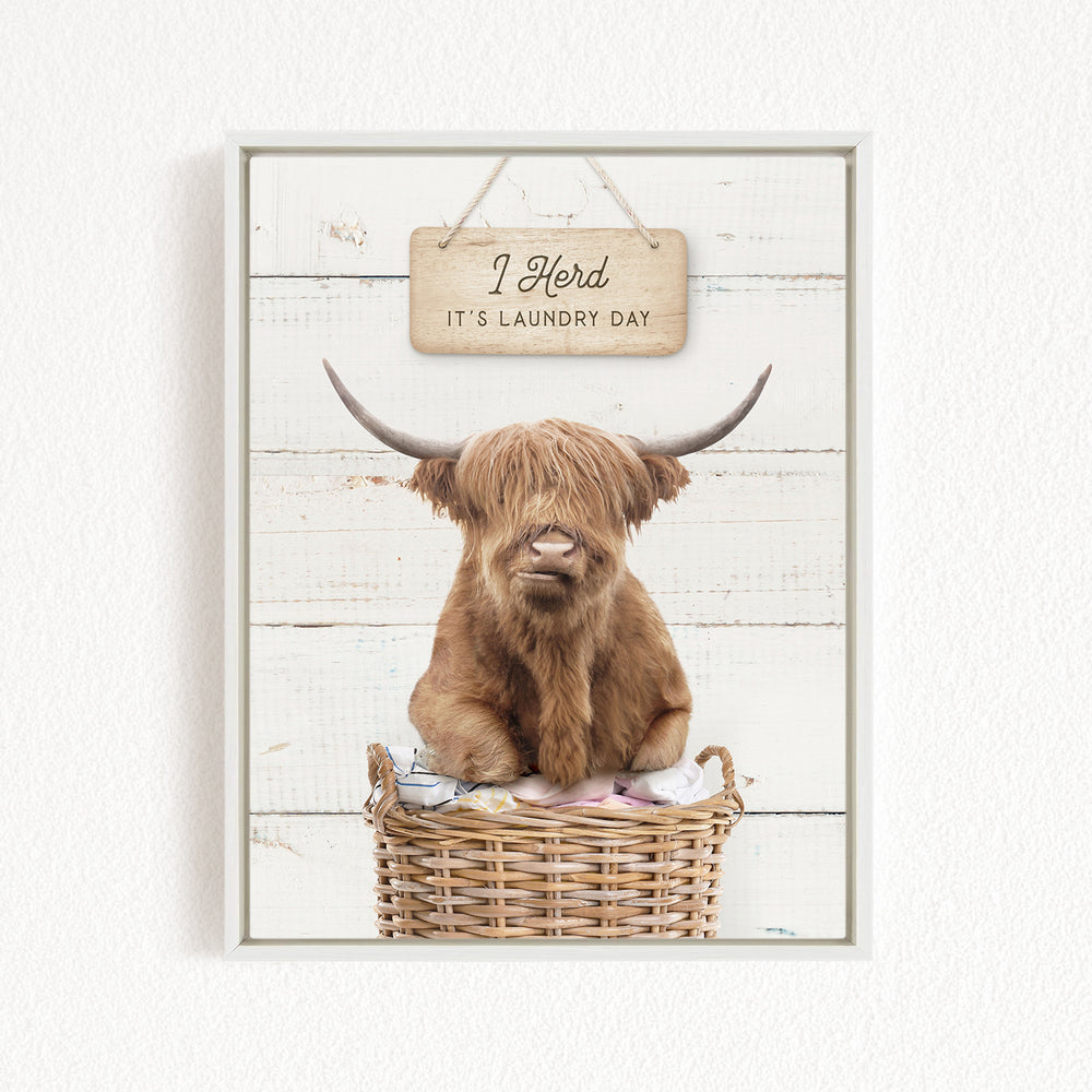 A brown cow with long horns is sitting in a wicker basket, with a sign above it that says "I herd it's laundry day".