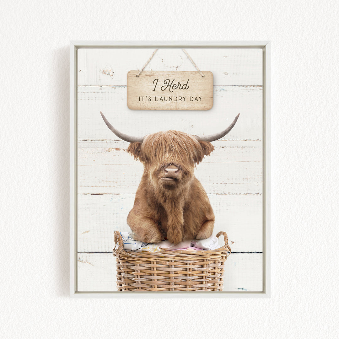 A brown cow with long horns is sitting in a wicker basket, with a sign above it that says "I herd it's laundry day".