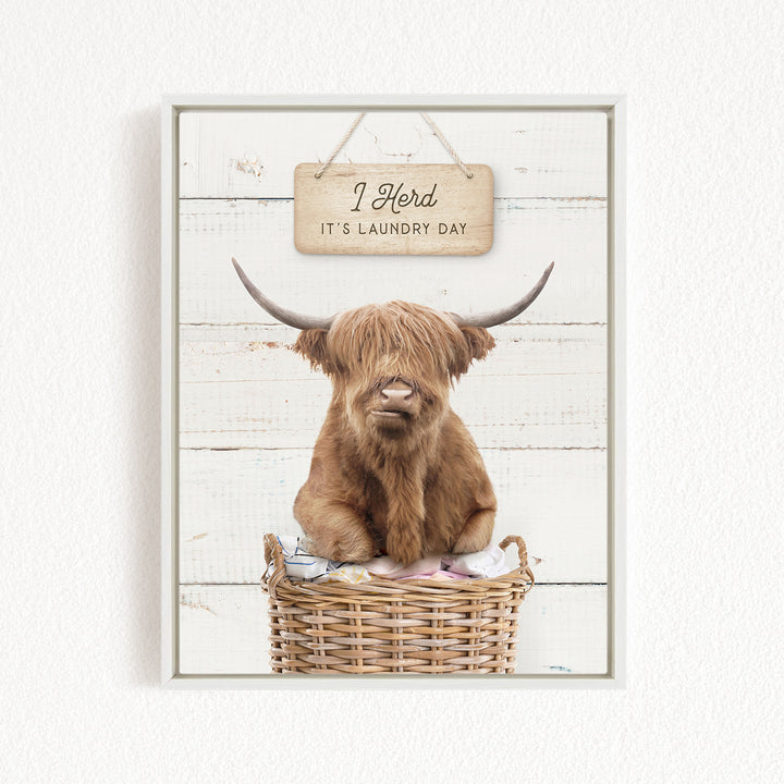 A brown cow with long horns is sitting in a wicker basket, with a sign above it that says "I herd it's laundry day".