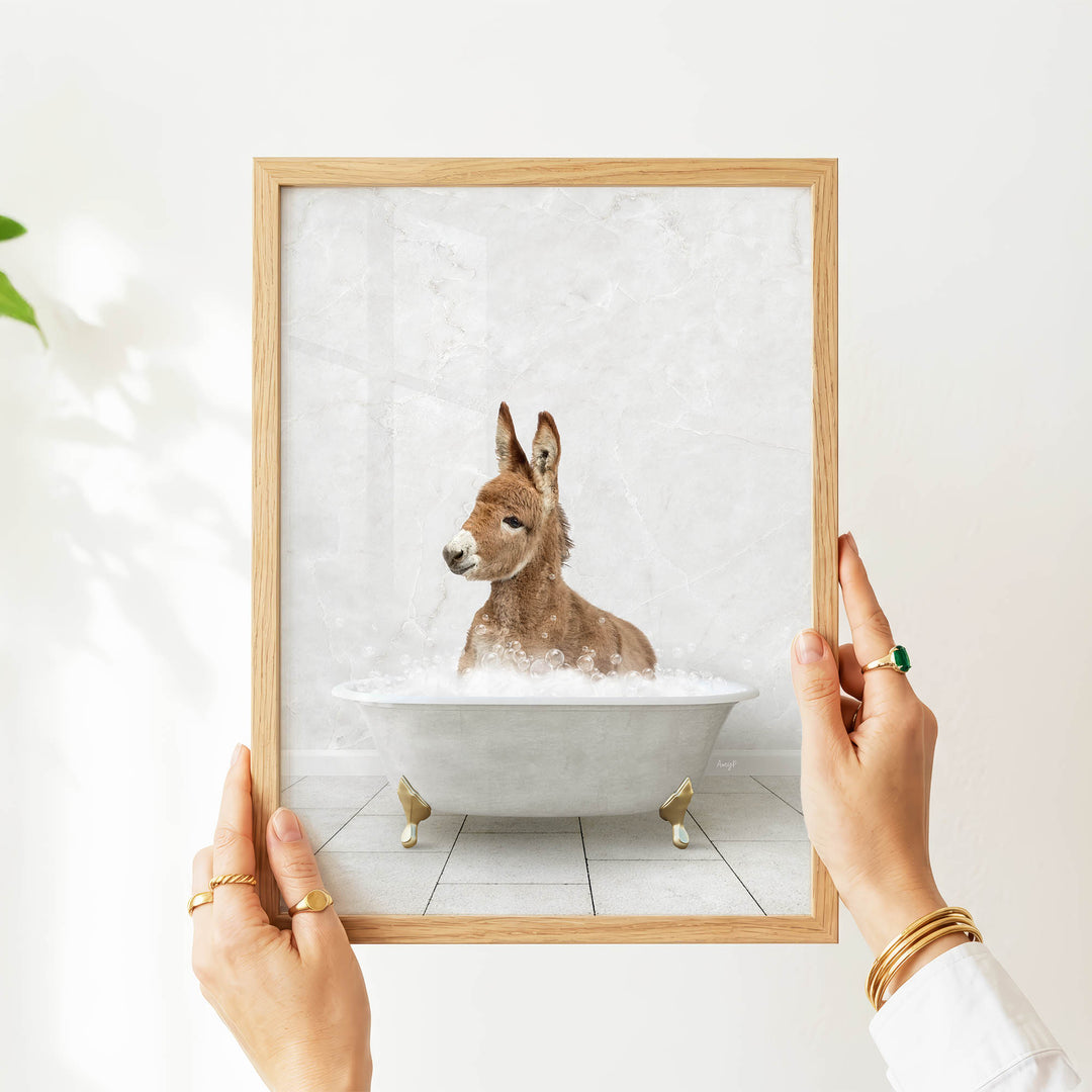 A person's hands are holding a framed picture of a donkey sitting in a bathtub filled with water.