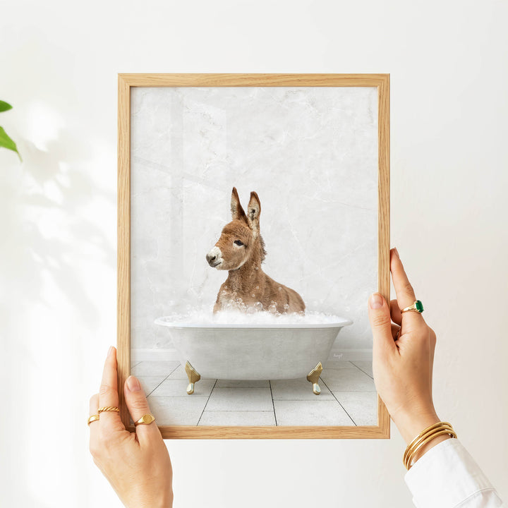 A person's hands are holding a framed picture of a donkey sitting in a bathtub filled with water.