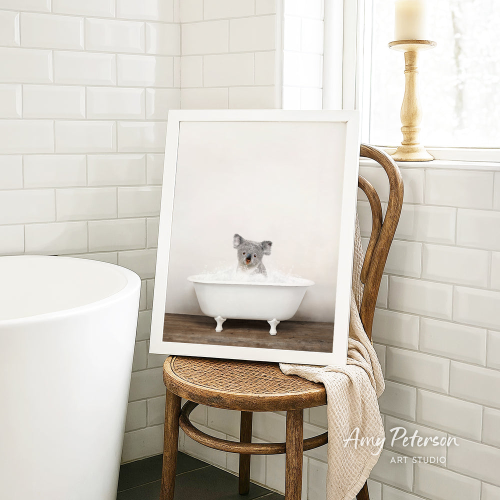 A framed photograph of a koala sitting in a bathtub, with a towel draped over the back of a wooden chair in a bathroom setting.