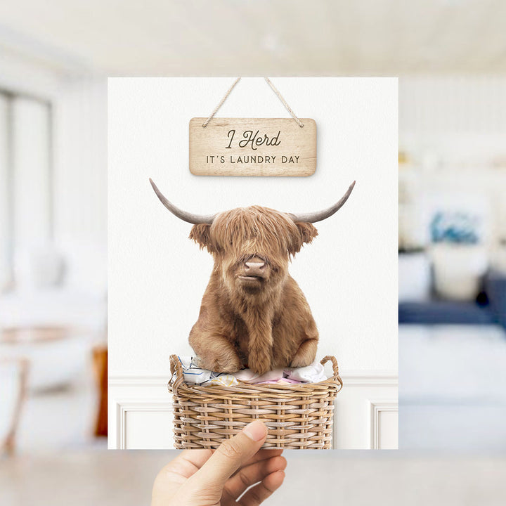 A hand holding a sign that says "I herd it's laundry day" above a wicker basket containing a large, horned animal, possibly a yak or a bull.