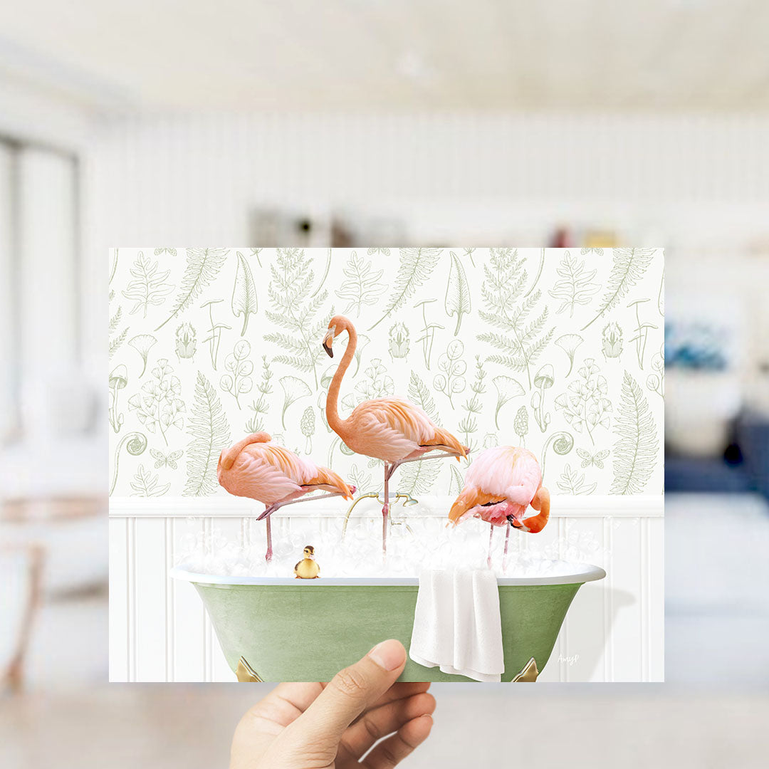 A hand holds up a framed image depicting three pink flamingos standing in a green bathtub filled with water and bubbles.