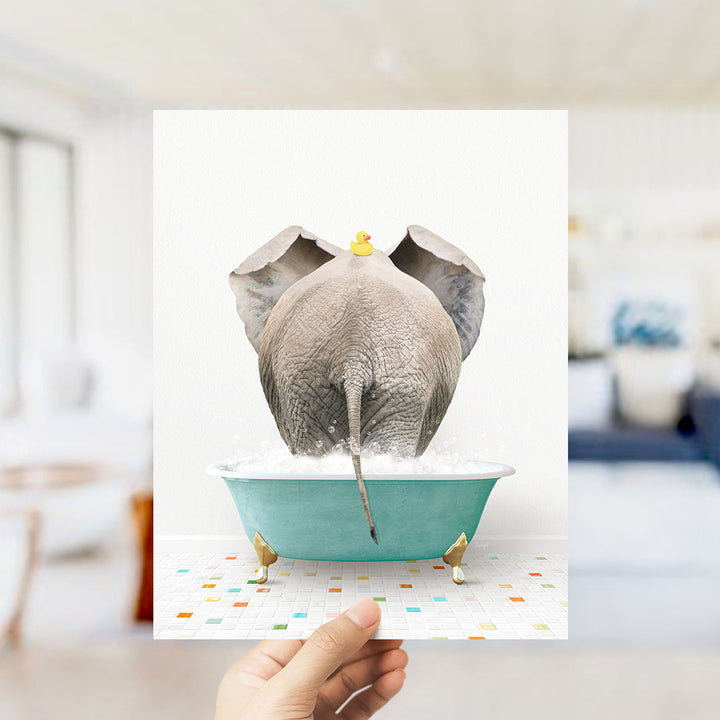 A hand holds up a framed image depicting an elephant sitting in a bathtub filled with bubbles, with a yellow rubber duck perched on its head.