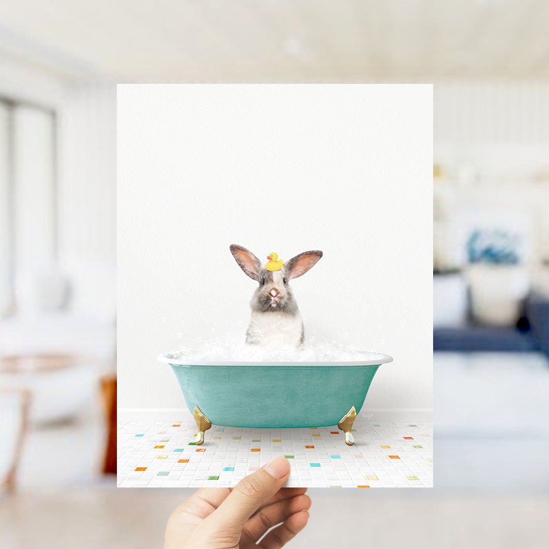 A hand holds up a framed image of a rabbit sitting in a bathtub filled with bubbles, wearing a yellow rubber duck.