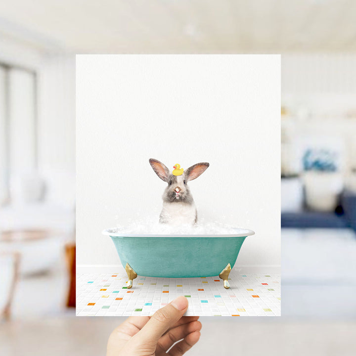 A hand holds up a framed image of a rabbit sitting in a bathtub filled with bubbles, wearing a yellow rubber duck.