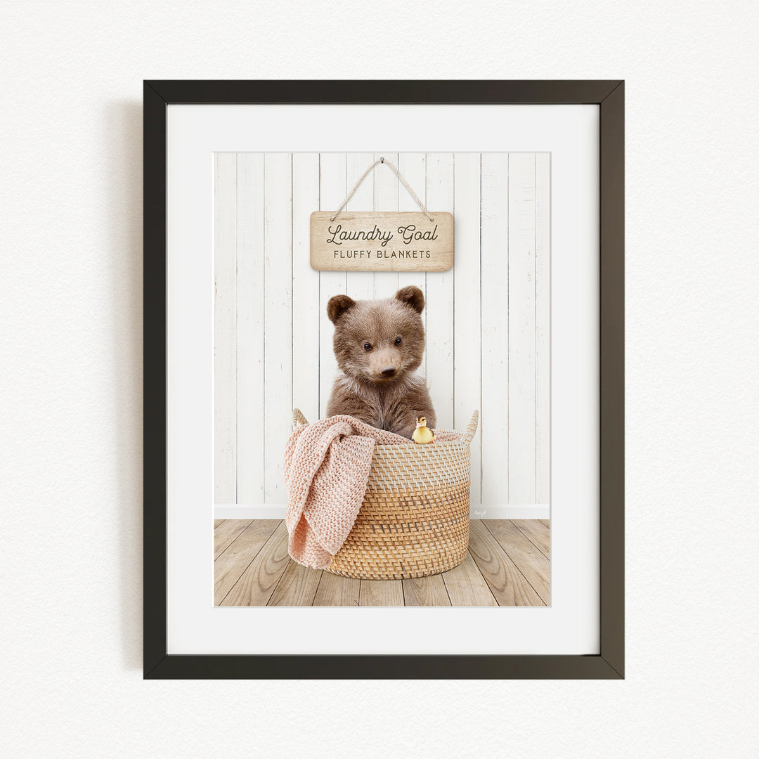 A framed image of a brown bear sitting in a woven basket, with a sign above the basket that reads "Laundry Goal Fluffy Blankets".