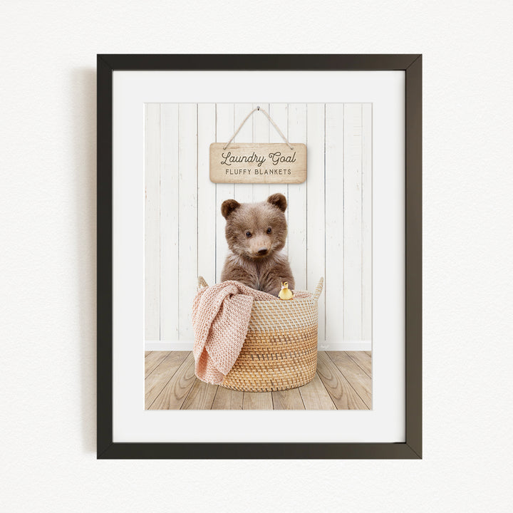 A framed image of a brown bear sitting in a woven basket, with a sign above the basket that reads "Laundry Goal Fluffy Blankets".
